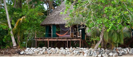 Una cabaña con techo de paja y una hamaca, rodeada por una costa rocosa y una exuberante vegetación tropical.