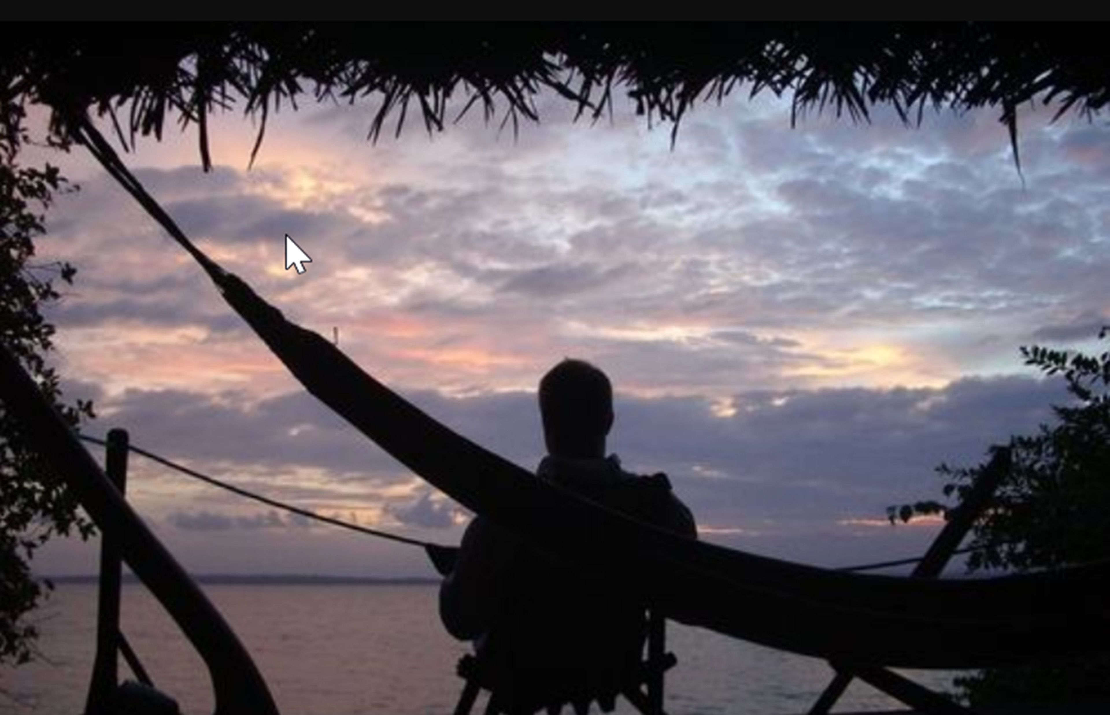 Uma rede com uma pessoa sentada, com vista para um corpo d'água e um céu ao pôr do sol.