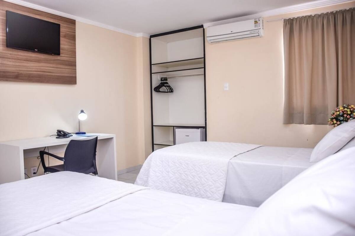 A hotel room with two beds, a desk with a chair, a TV mounted on the wall, an air conditioning unit, and a window with curtains.
