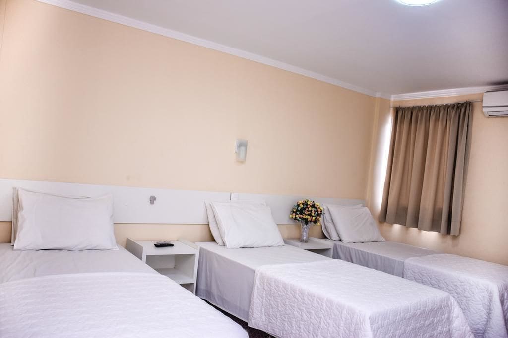 Une chambre d’hôtel avec trois lits simples, une table de chevet avec une lampe, un vase avec des fleurs et une fenêtre avec des rideaux.