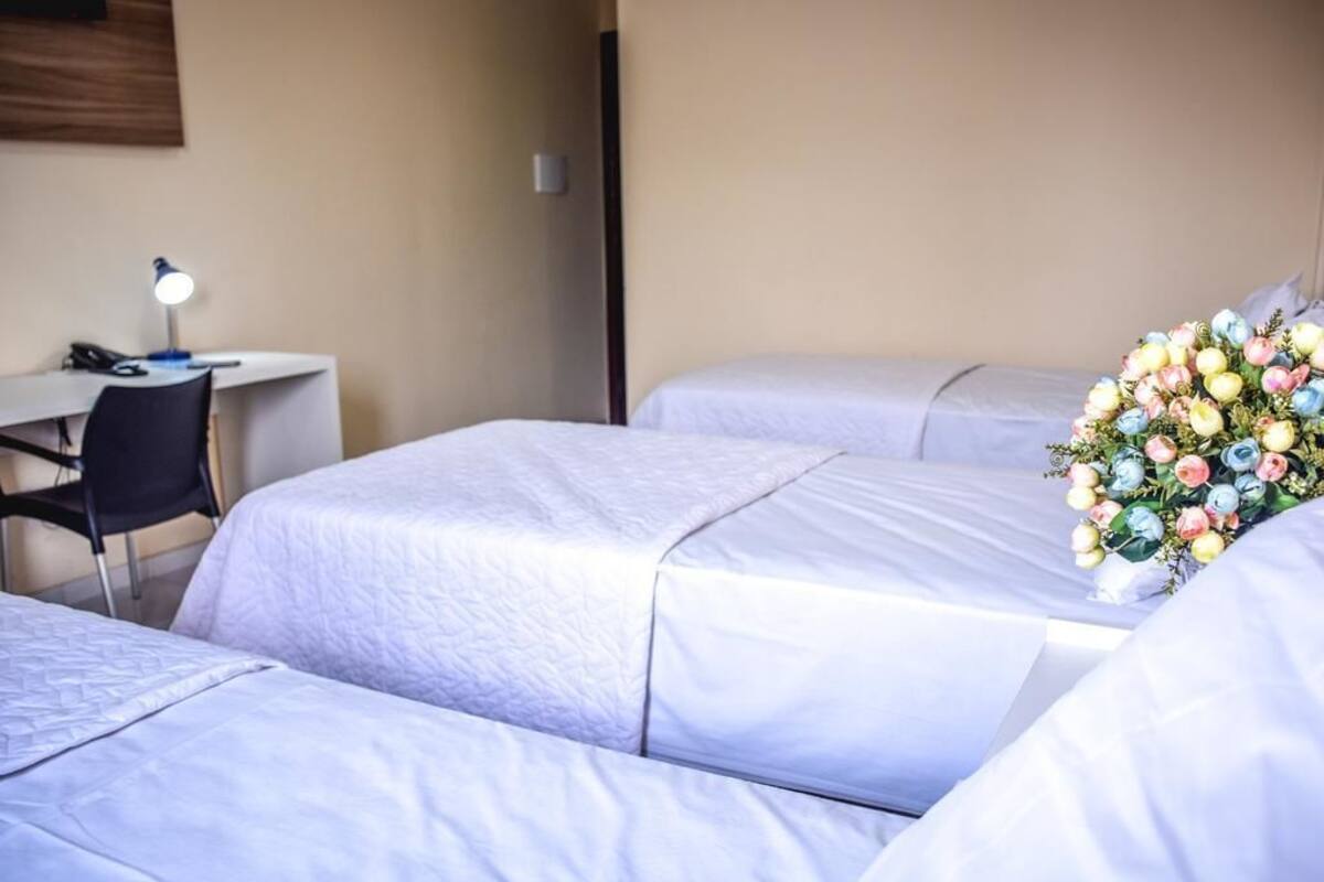 A hotel room with three beds, a desk with a lamp, and a floral arrangement.