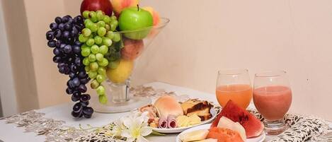 Una mesa con un frutero que contiene uvas, manzanas, melocotones, dos vasos de jugo y platos con sándwiches y una rebanada de sandía.