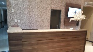 A reception desk with a wooden finish, a patterned wall behind it, and a television mounted above. - Prime Hotel (Feira de Santana)