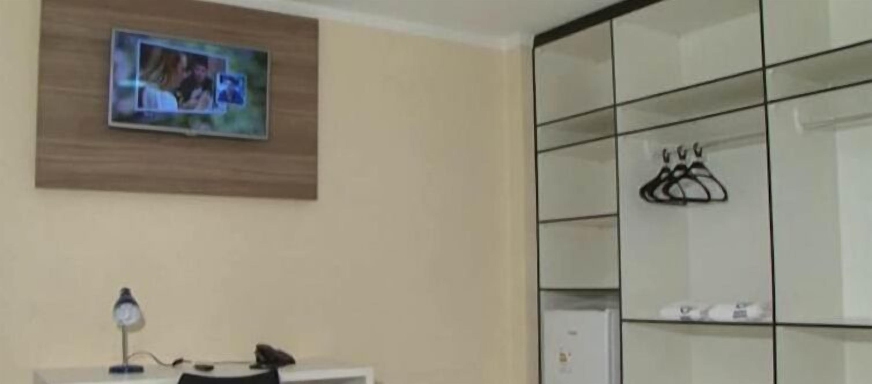 Una habitación con televisor montado en la pared, un escritorio con lámpara y un armario con espejo y perchas.