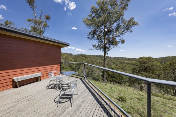 Una terraza de madera con una mesa y sillas de metal, un banco y una barandilla con vistas a un paisaje montañoso.