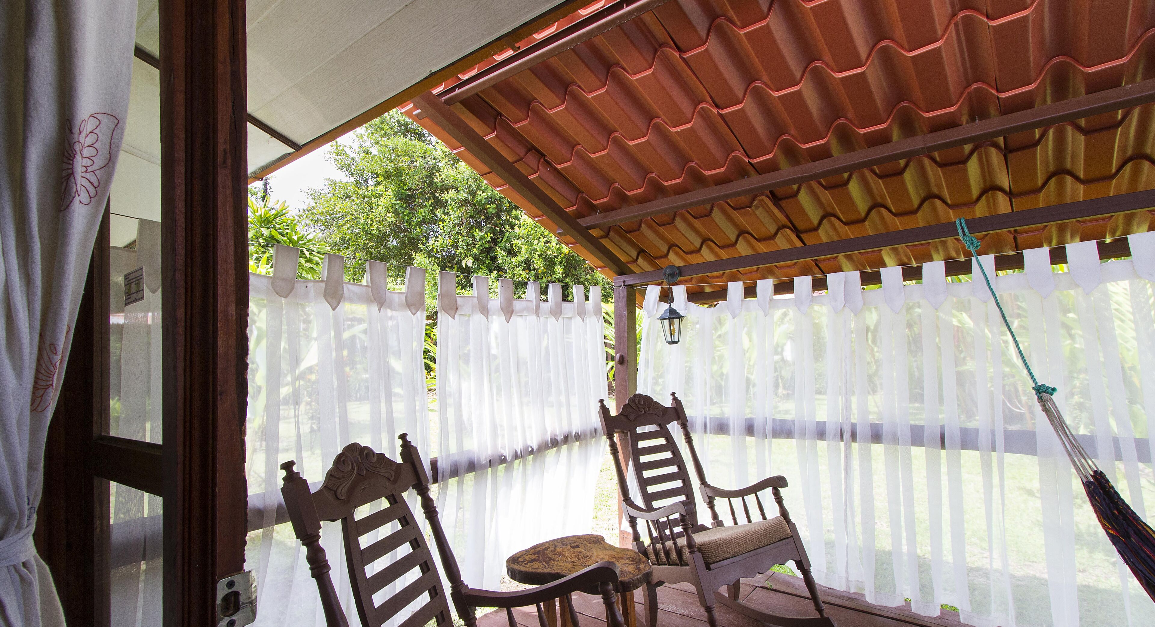 Un patio techado con dos sillas de madera y una mesa pequeña, rodeado por una cerca blanca y un techo rojo.