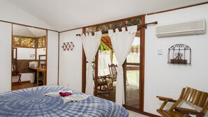 Um quarto com cama, uma cadeira de madeira, uma mesinha e uma janela com cortinas.