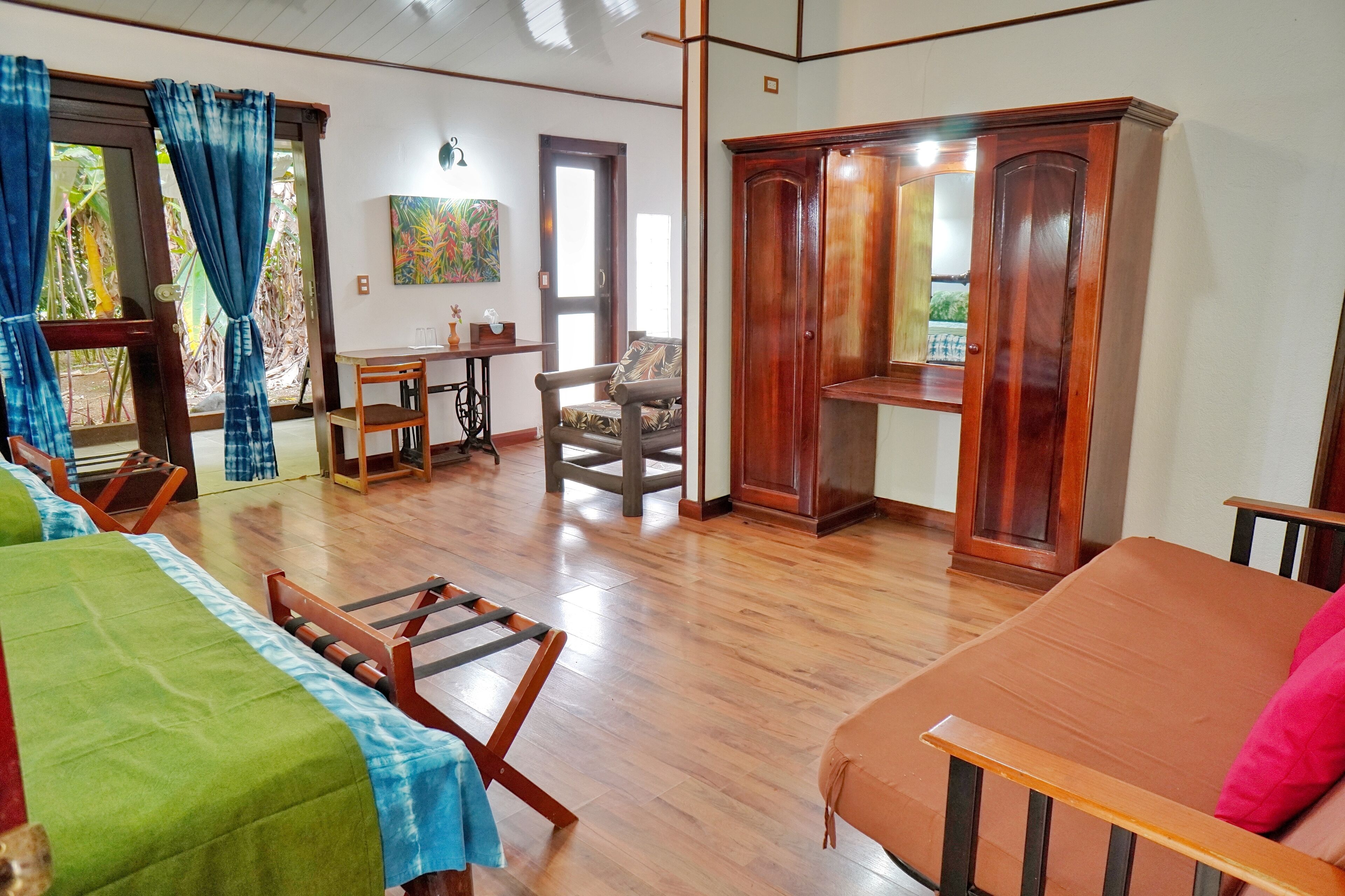A room with wooden flooring, a bed with a green and blue bedspread, a wooden chair, a wooden table, and a wardrobe.