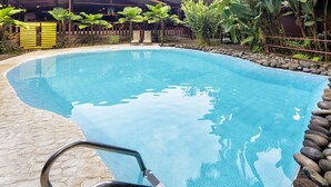 Piscina azul límpida com uma escada de metal, cercada por uma borda de pedra e plantas tropicais.