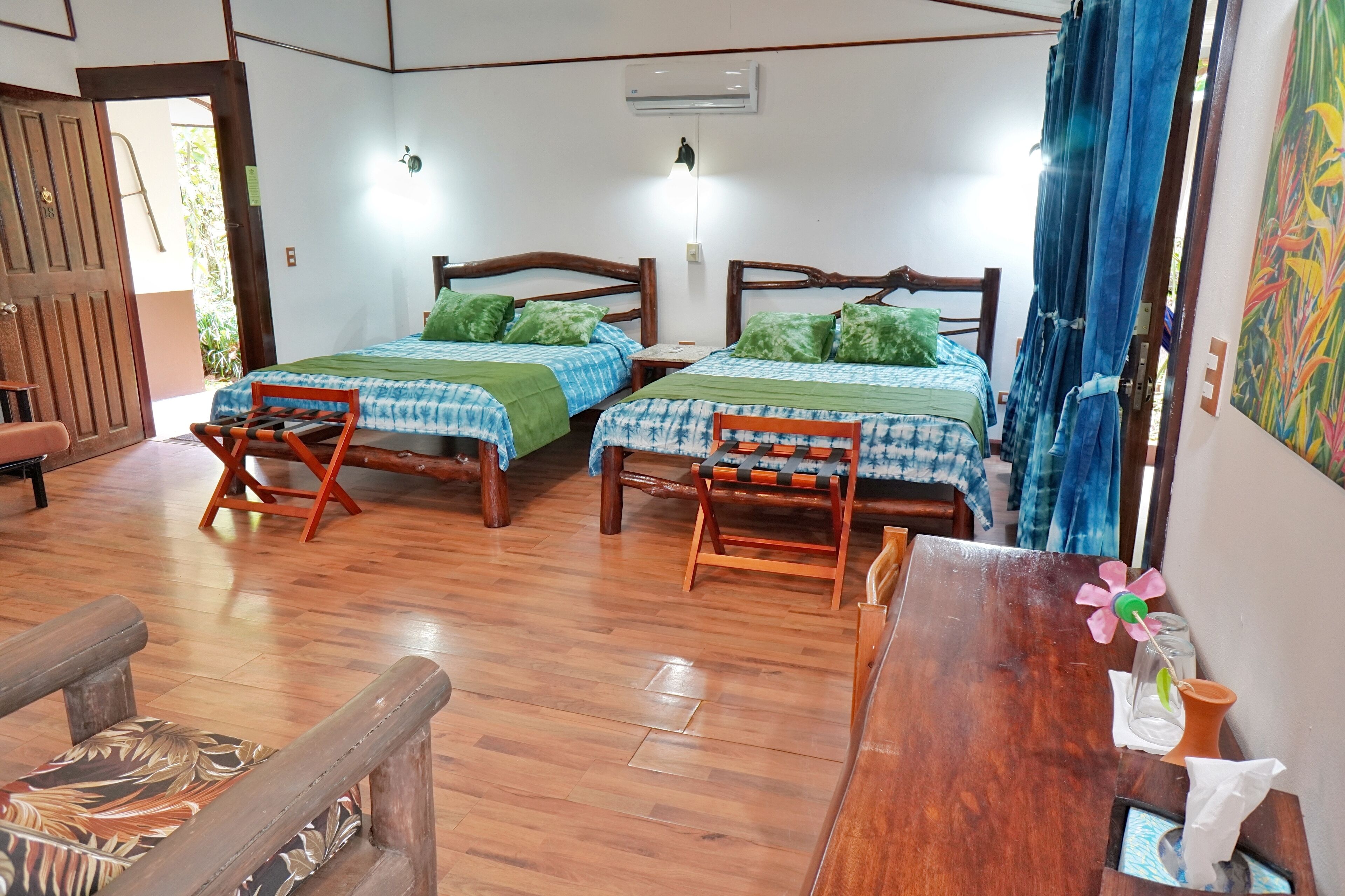 A room with two wooden beds, each with a green bedspread and pillows, a wooden table with a vase and a flower, and a wooden bench.