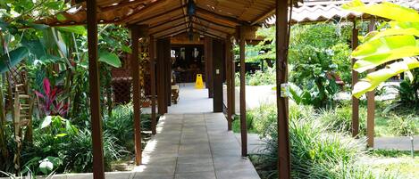 A covered walkway with a tiled floor, surrounded by lush greenery and plants.