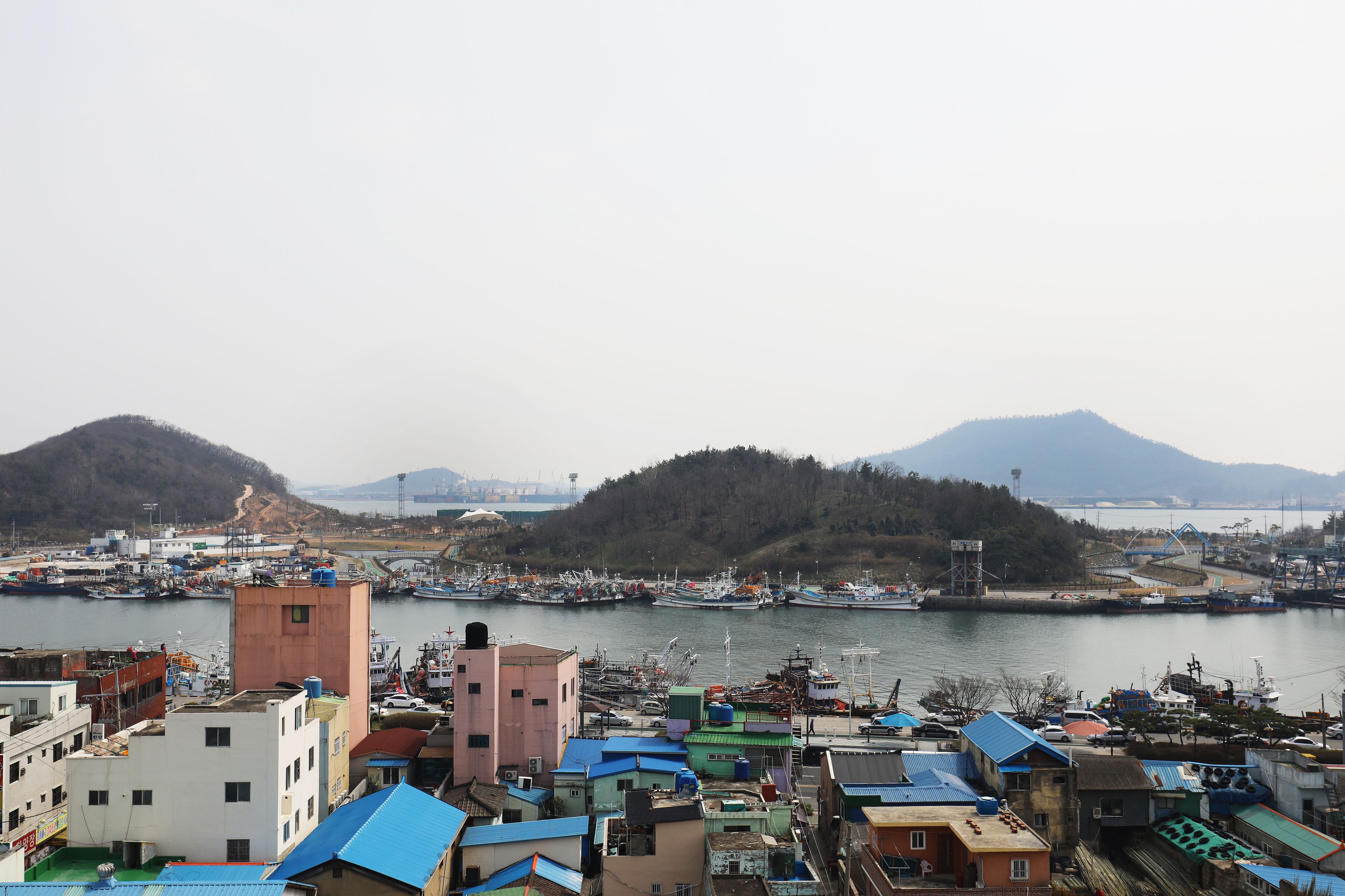 Un pueblo costero con un puerto deportivo, edificios con techos azules y un paisaje montañoso al fondo.