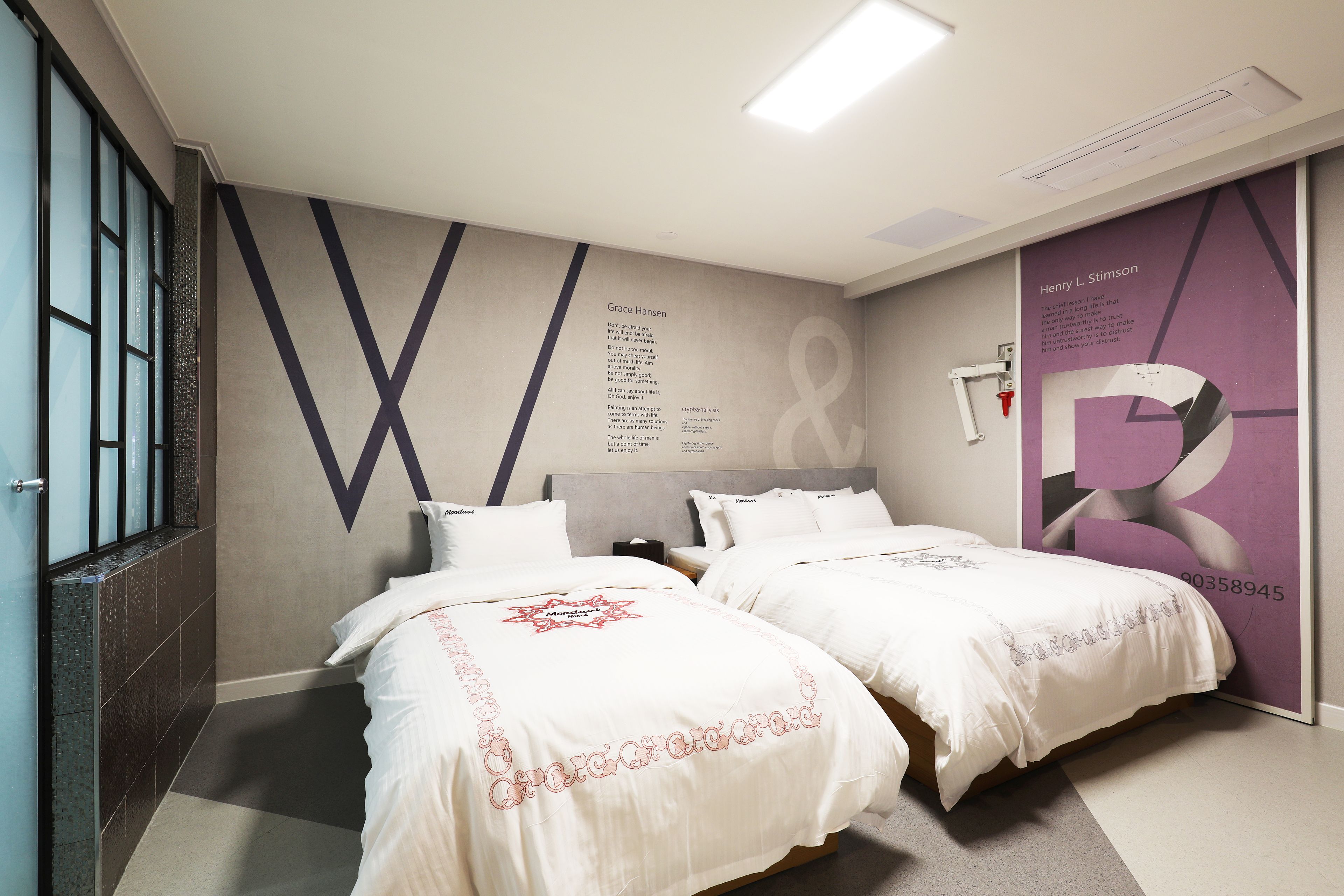 Ein Hotelzimmer mit zwei Betten, einem großen Fenster und einer Wand mit einem geometrischen Muster und Text.