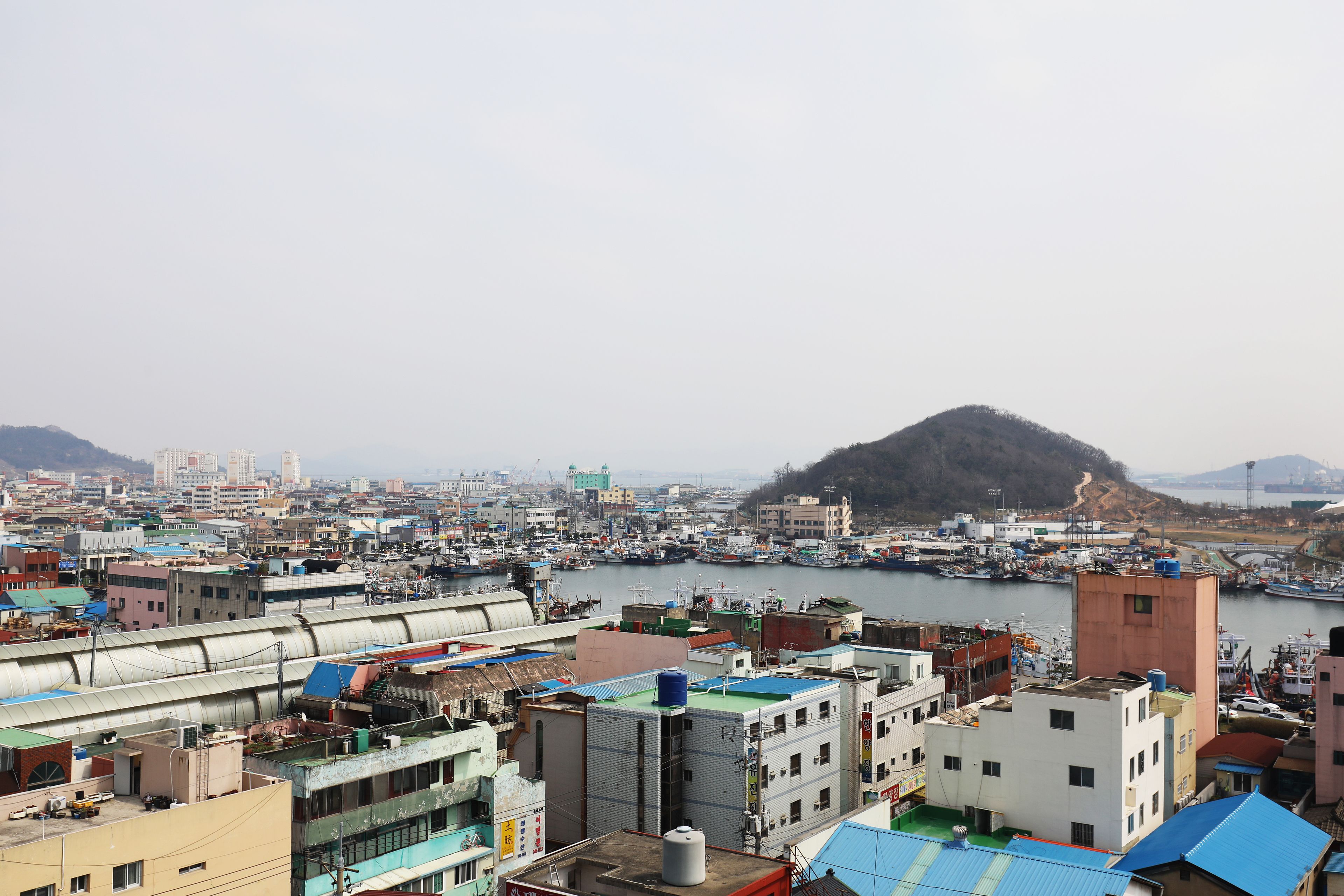 A coastal cityscape with a marina, industrial buildings, and a hill in the background.