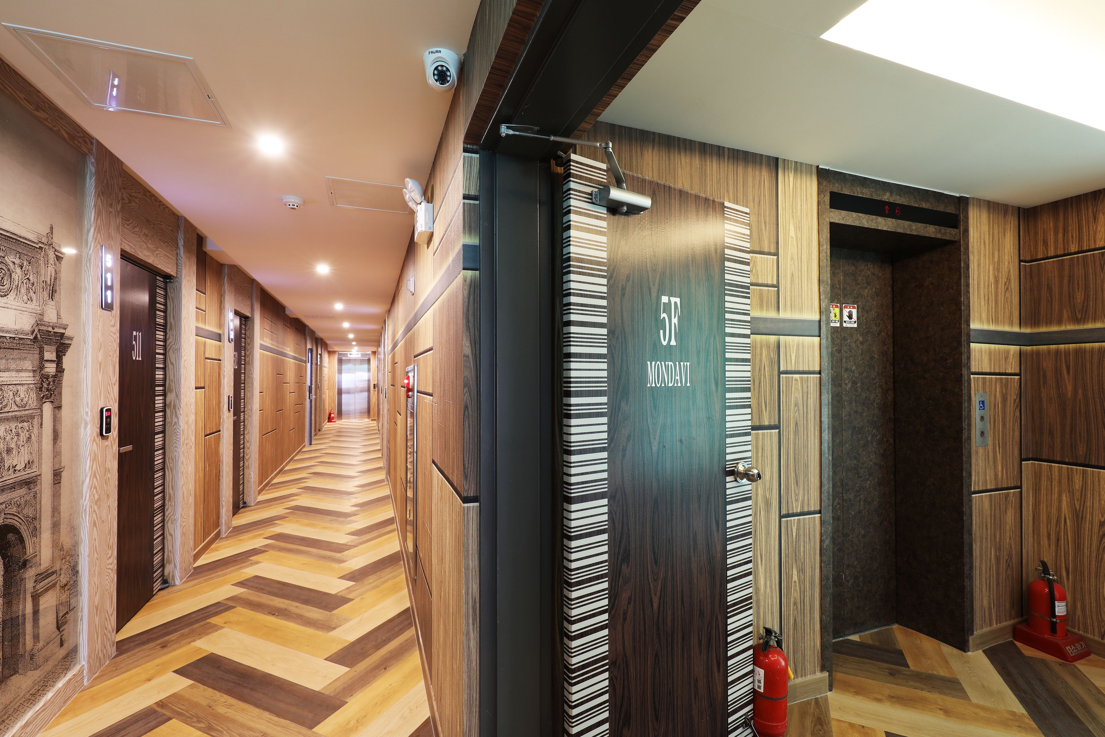 A hotel corridor with wooden paneling, a herringbone floor pattern, and a fire extinguisher near the elevator.