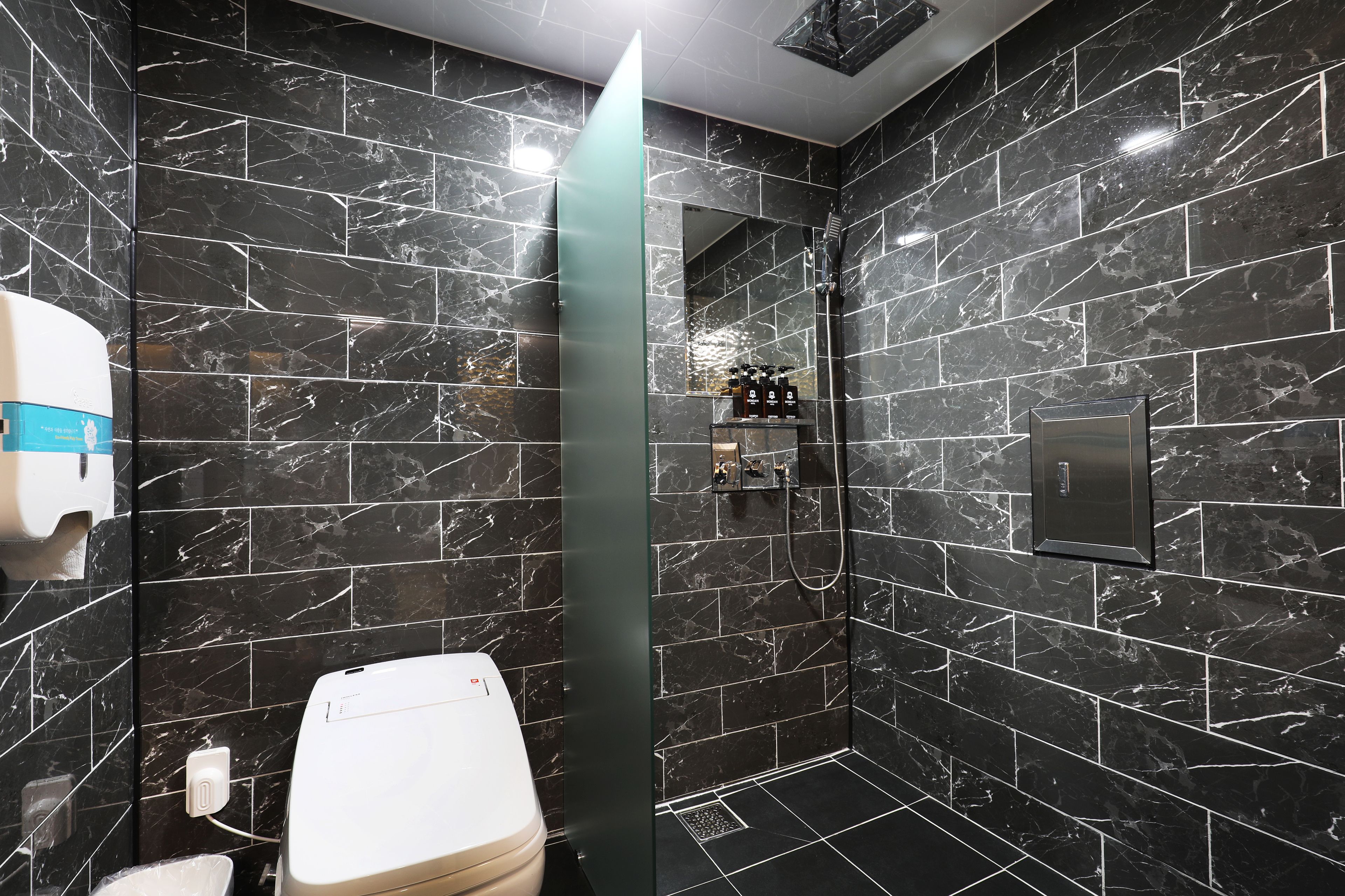 A modern bathroom with black marble tiles, a glass shower enclosure, and a toilet with a paper towel dispenser.