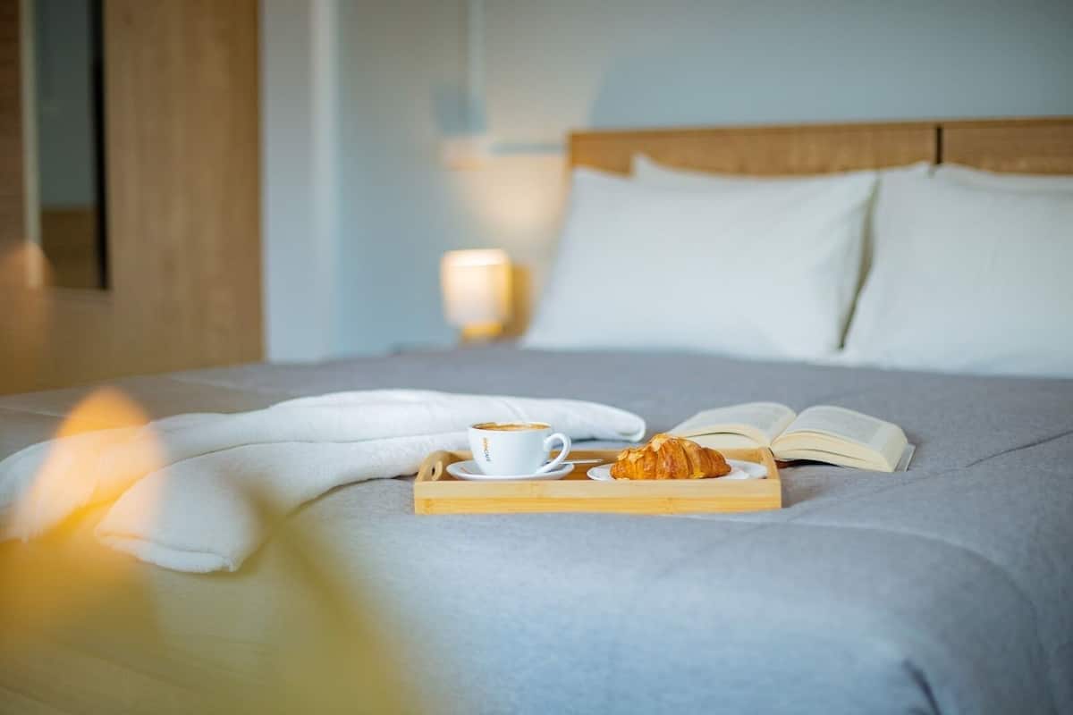 Una cama bien hecha con una bandeja de croissant y café, un libro abierto y una lámpara de noche.