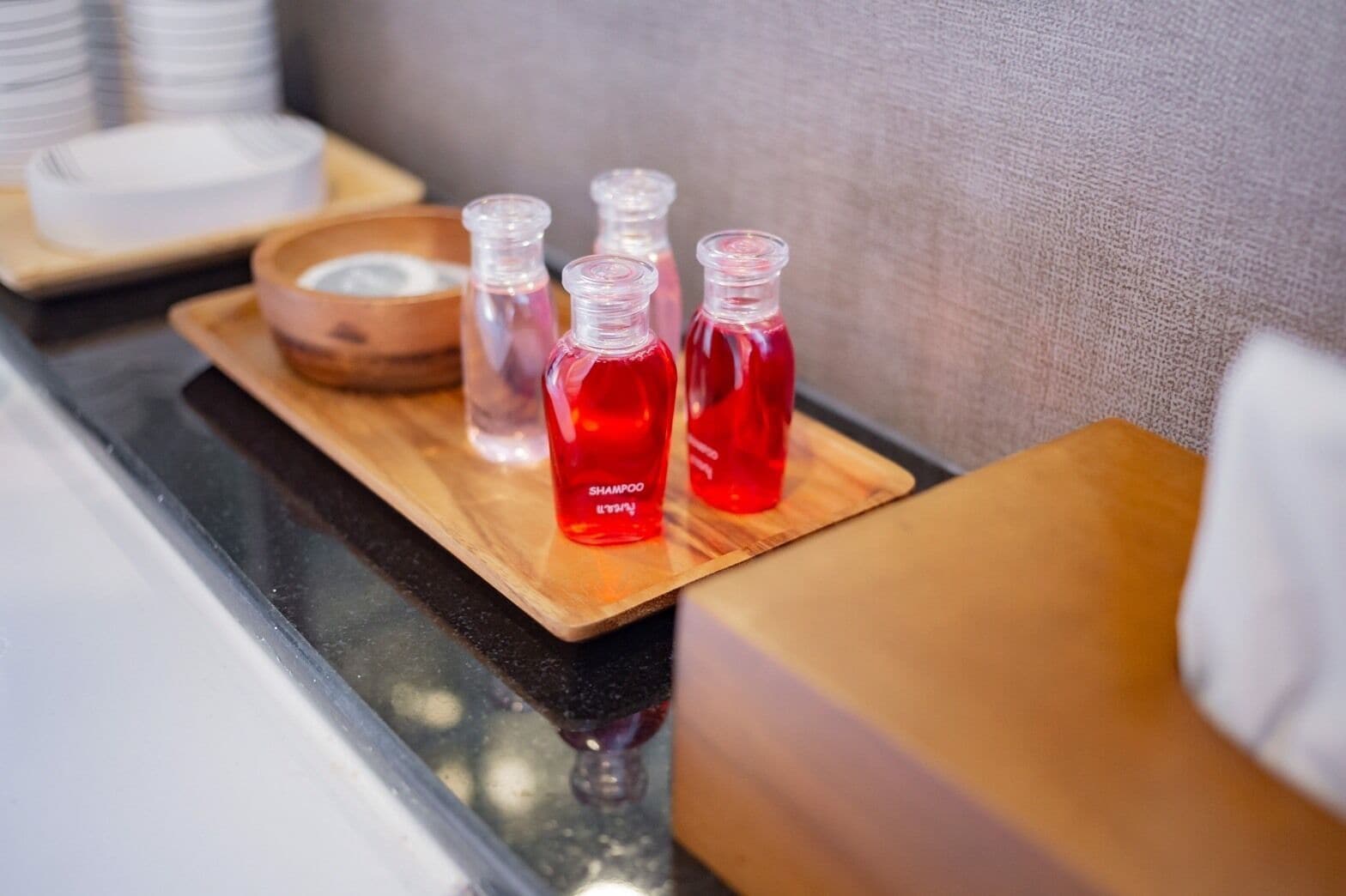 A bathroom counter with a wooden tray holding four bottles of red shampoo and a bowl of white soap.