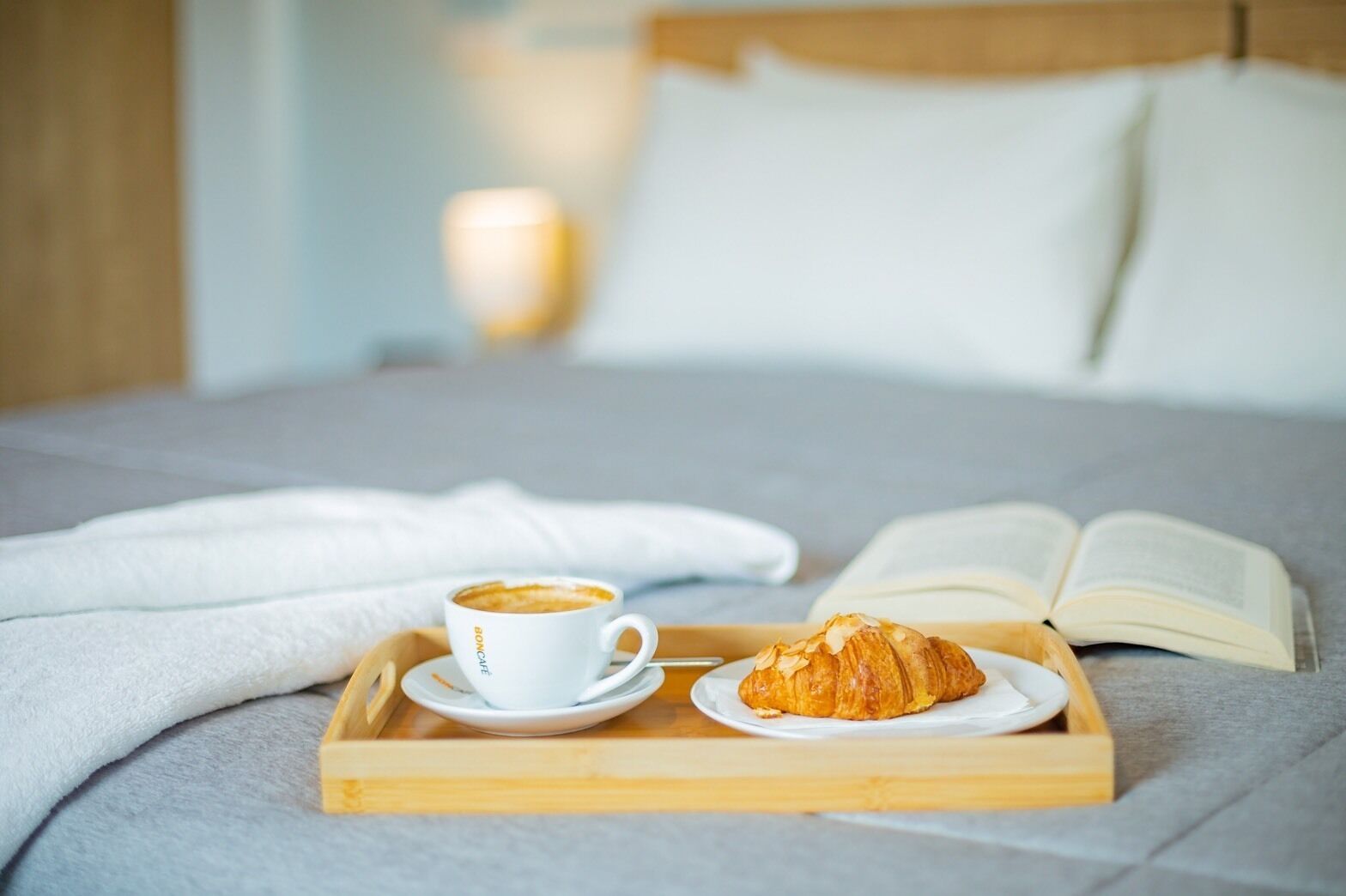 A bed with a tray containing a croissant and a cup of coffee, an open book, and a bedside lamp.