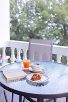 Una mesa redonda de vidrio con un libro, un croissant, un vaso de jugo de naranja y una taza de café en un balcón con vista a árboles.