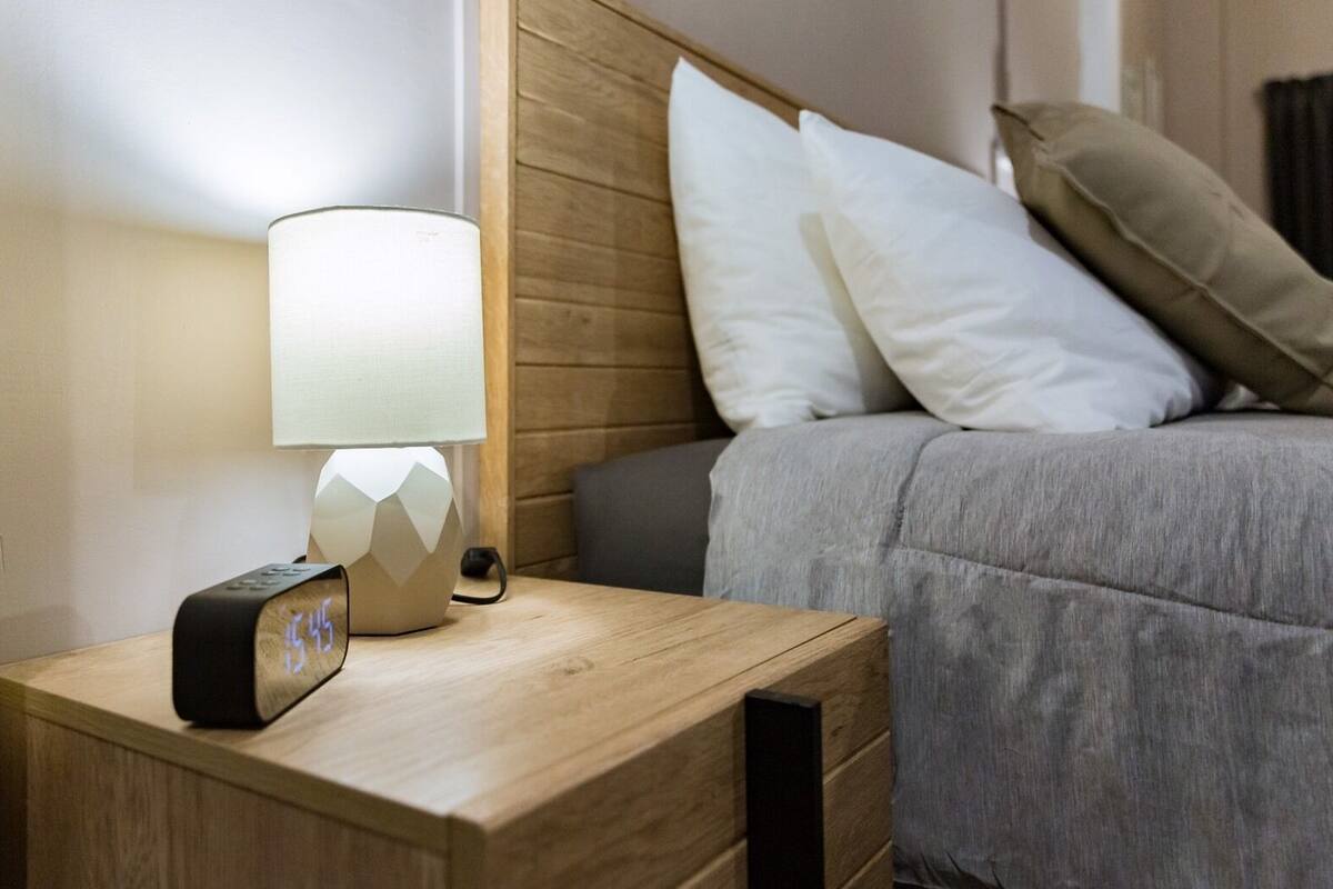 Una cama bien hecha con edredón gris, cabecera de madera y mesita de noche con lámpara y reloj digital.