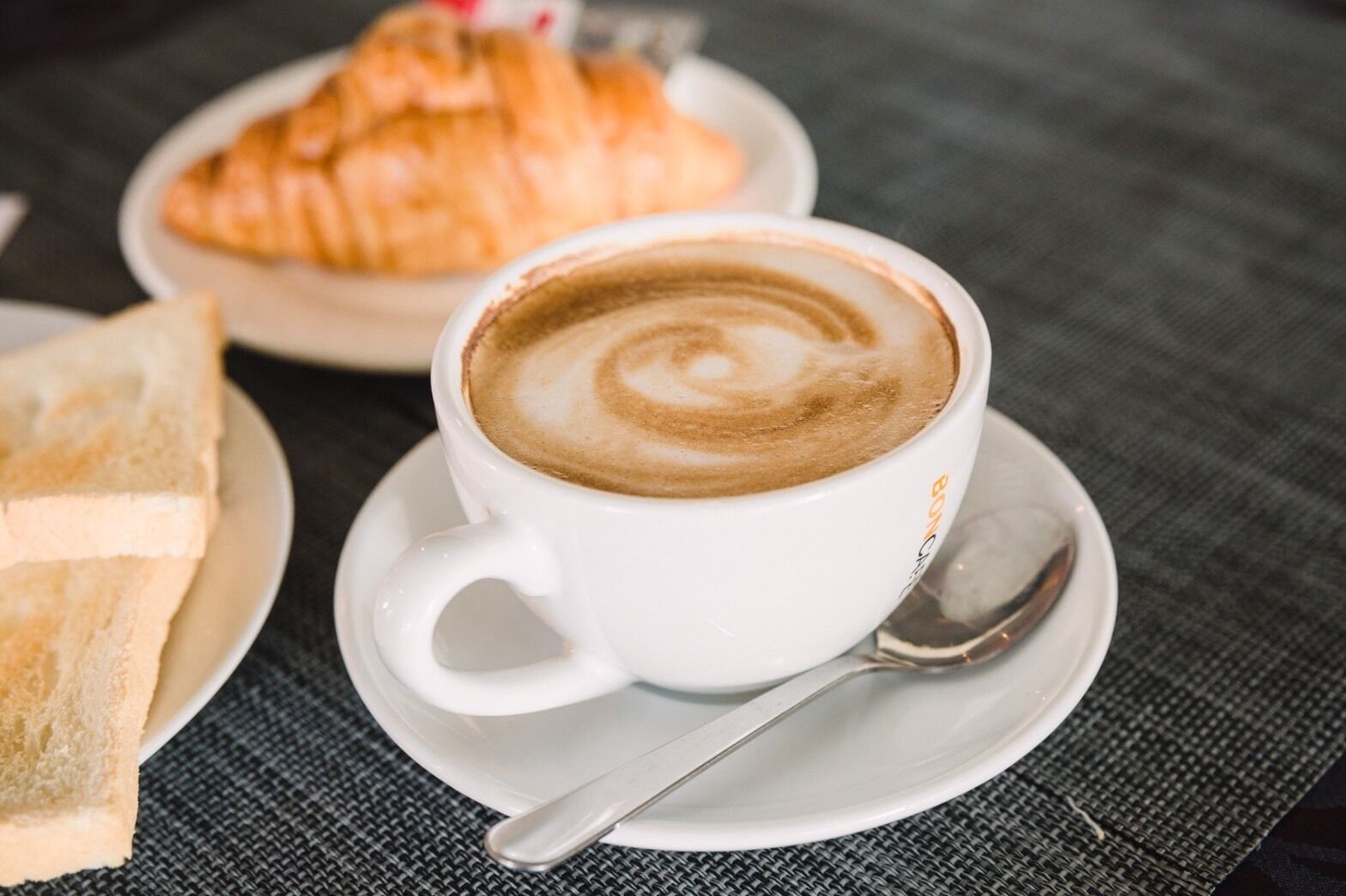A cup of coffee with latte art on a saucer, accompanied by a croissant and slices of bread on a table.