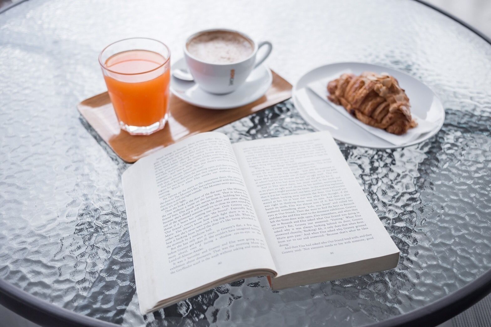 An open book on a glass table with a glass of orange juice and a cup of coffee.
