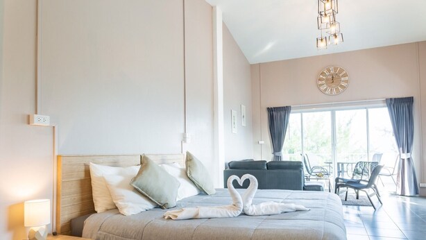 A modern hotel room with a large bed, a wooden headboard, a bedside table with a lamp, and a view of the outdoor seating area. - The Beach Cha Am Suite (Cha-am)
