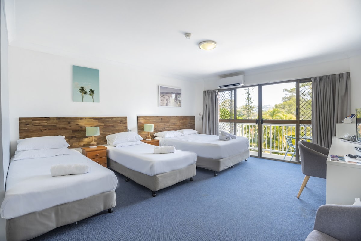A hotel room with three beds, a desk, and a view of the outdoors.