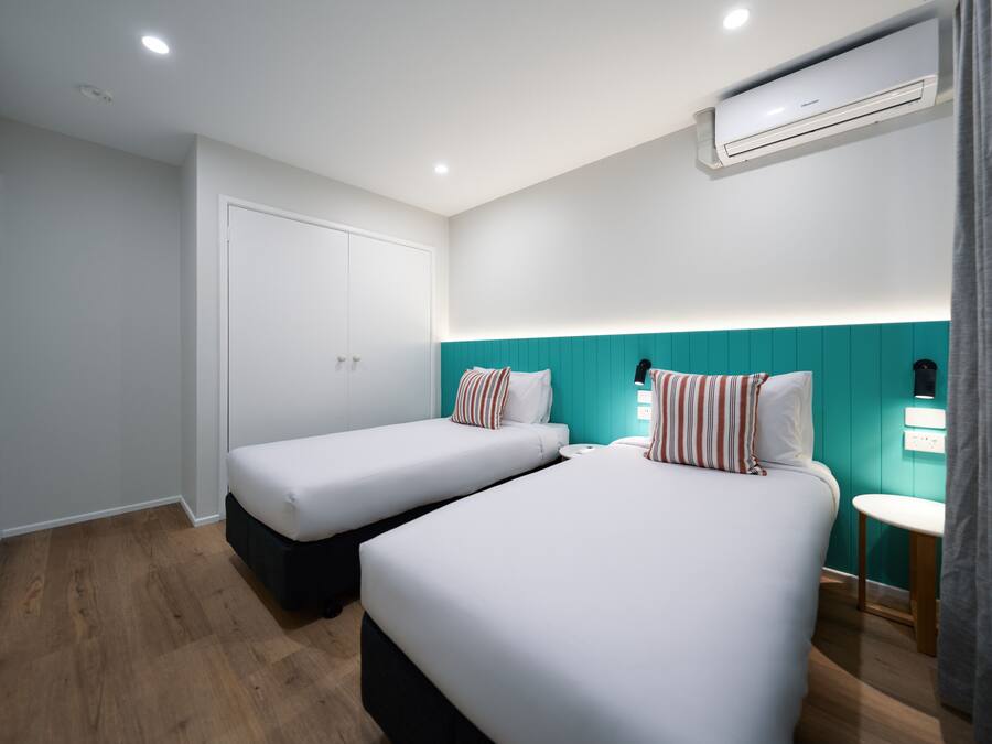 A hotel room with two beds, a white door, a teal accent wall, an air conditioning unit, and a small bedside table.