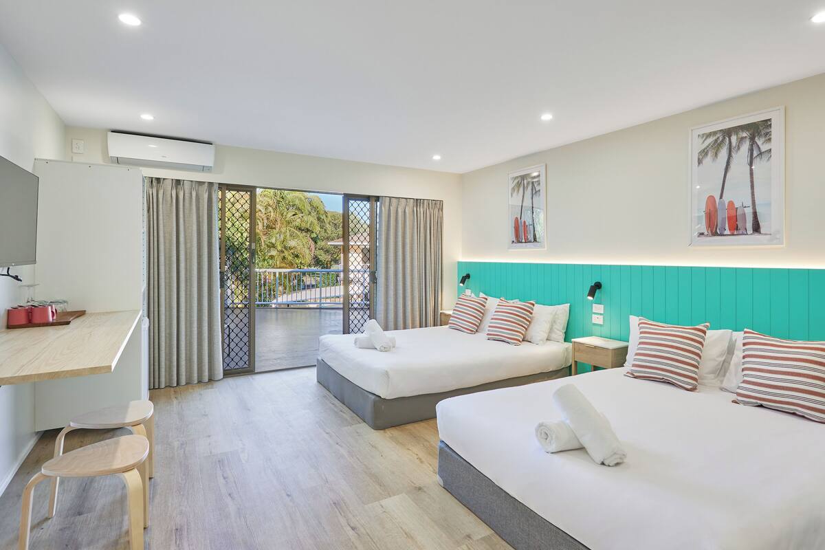 A hotel room with two beds, a desk, and a view of a pool and palm trees.