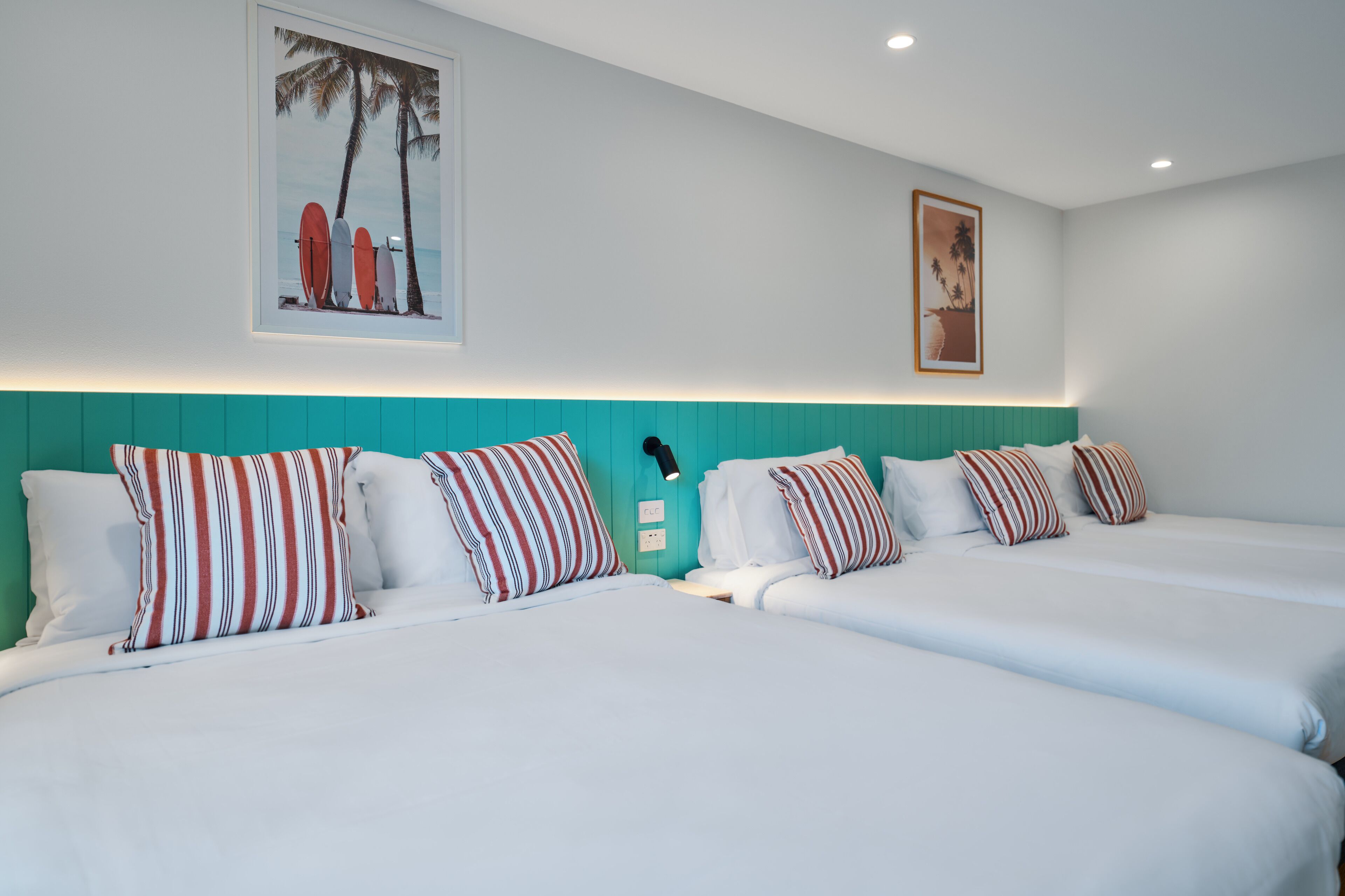 Habitación de hotel con dos camas, cabecero verde azulado, almohadas a rayas y dos cuadros en la pared.