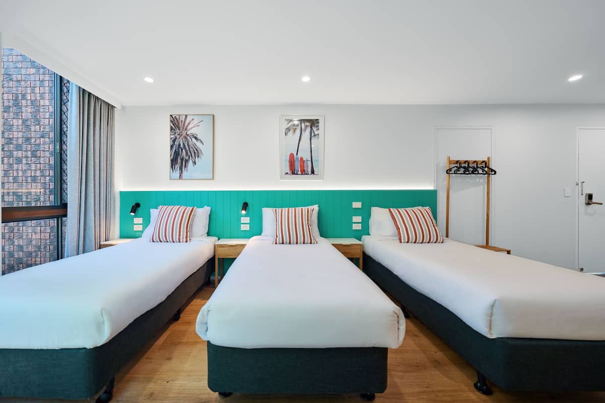 A hotel room with three single beds, teal-colored headboards, white bedding, and two wall-mounted artworks.