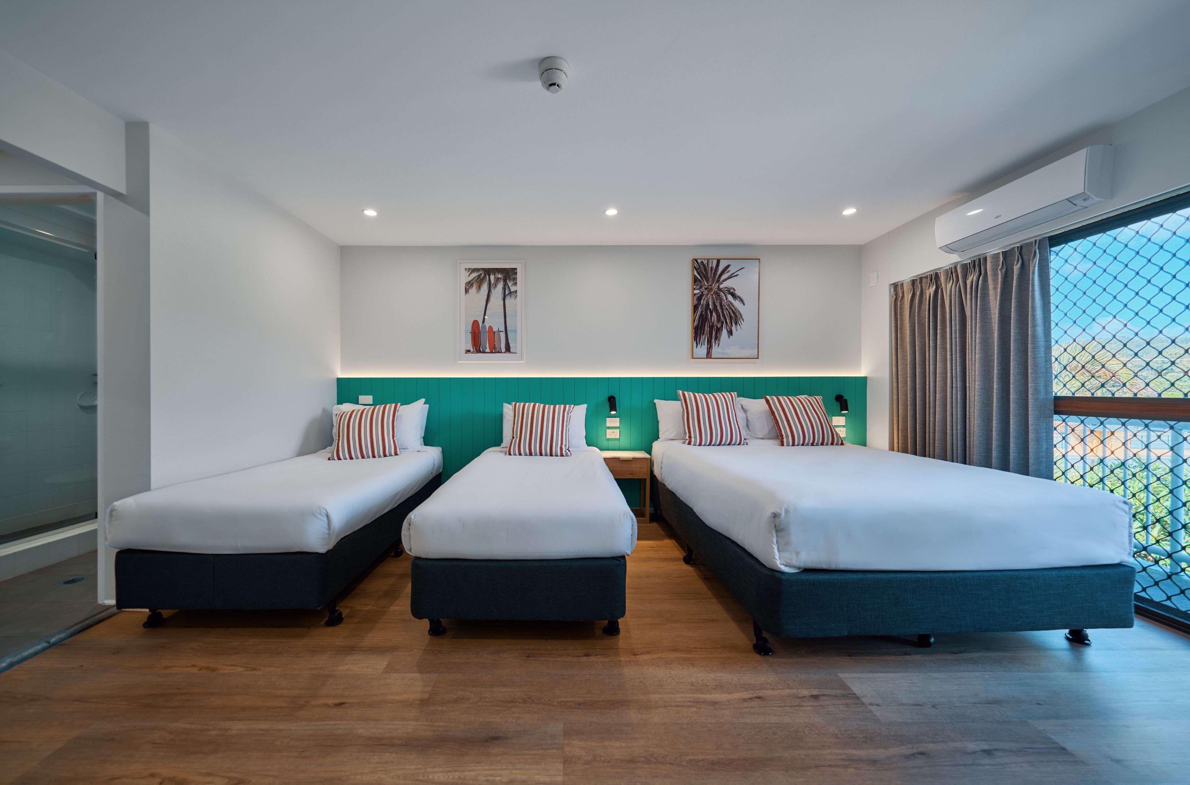 A hotel room with three single beds, a green accent wall, and a large window with a view.
