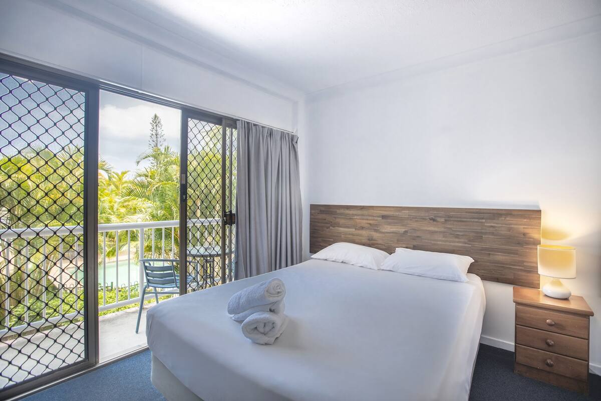 A hotel room with a bed, a nightstand, a lamp, a sliding glass door to a balcony, and a view of greenery.