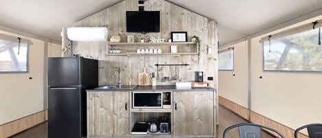 A compact kitchen with a built-in refrigerator, a sink, and wooden cabinetry. There is a flat-screen TV mounted on the wall, and a window providing natural light.