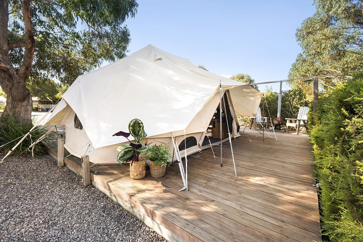 A large white tent with a wooden deck, surrounded by trees and potted plants.