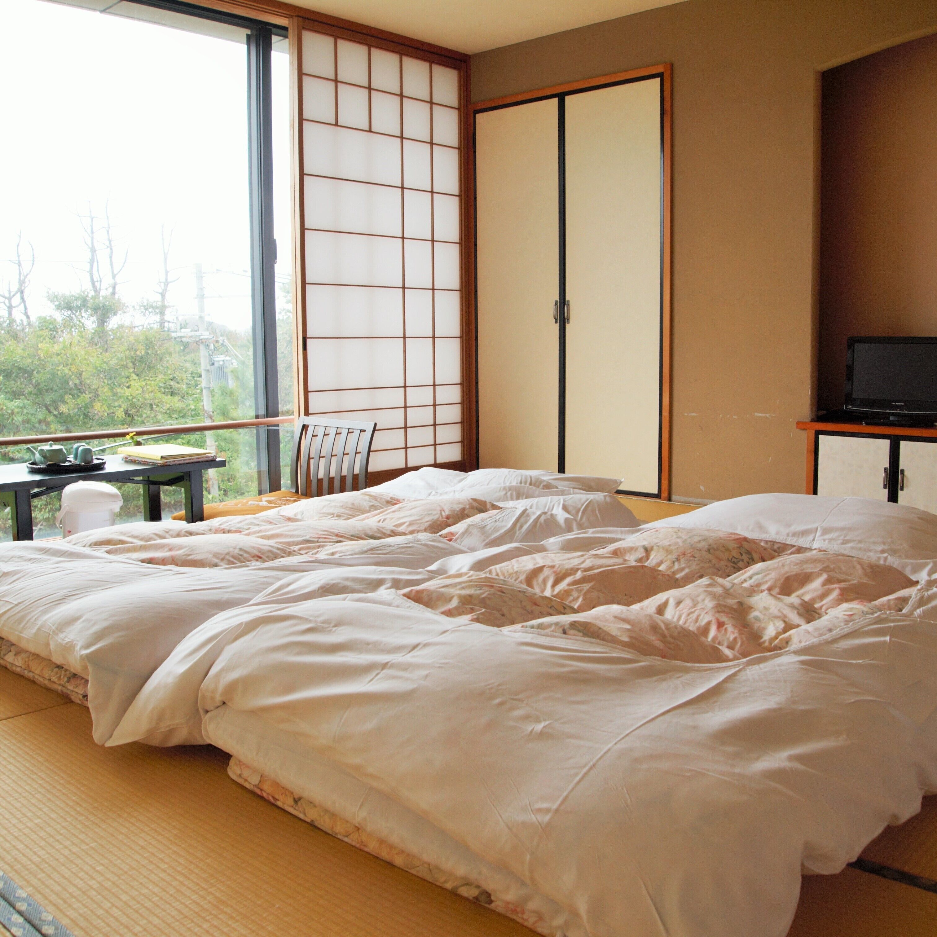 A bedroom with a large bed, a futon, a sliding door, a TV, and a view of greenery outside.