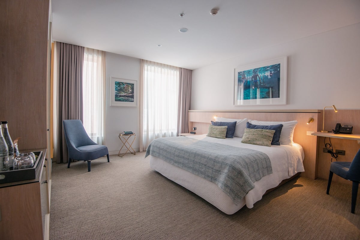 A hotel room with a large bed, a chair, a desk, and a painting on the wall.