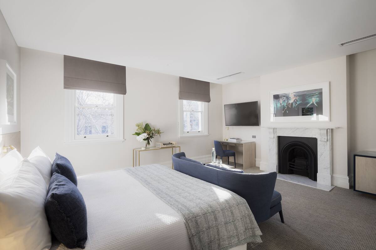 A modern hotel room with a large bed, a desk, a sofa, a fireplace, and a TV.
