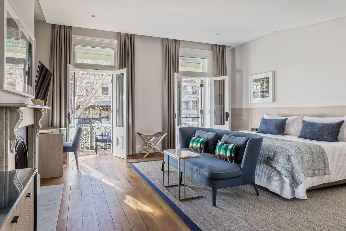 A modern hotel room with a large bed, a sofa, a small table, and a view of the city through large windows.