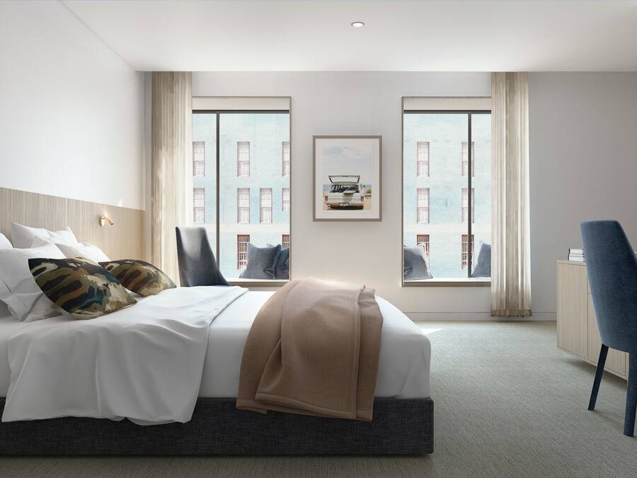Une chambre d’hôtel moderne, équipée d’un lit, d’un bureau et d’une chaise. Les fenêtres offrent une vue sur les bâtiments environnants.
