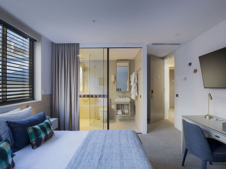 Habitación de hotel moderna con una cama grande, un escritorio, un televisor y un baño visible a través de una puerta de cristal.