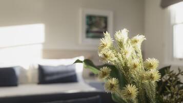Uma cama com roupa de cama branca e almofadas escuras, um vaso com flores brancas e um quadro na parede.