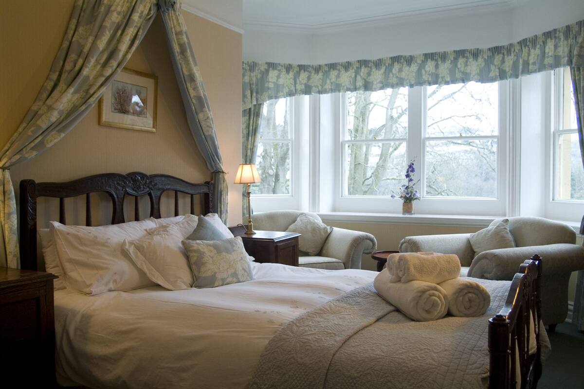 Une chambre à coucher avec un grand lit, deux fauteuils, une table de chevet et une fenêtre avec des rideaux.
