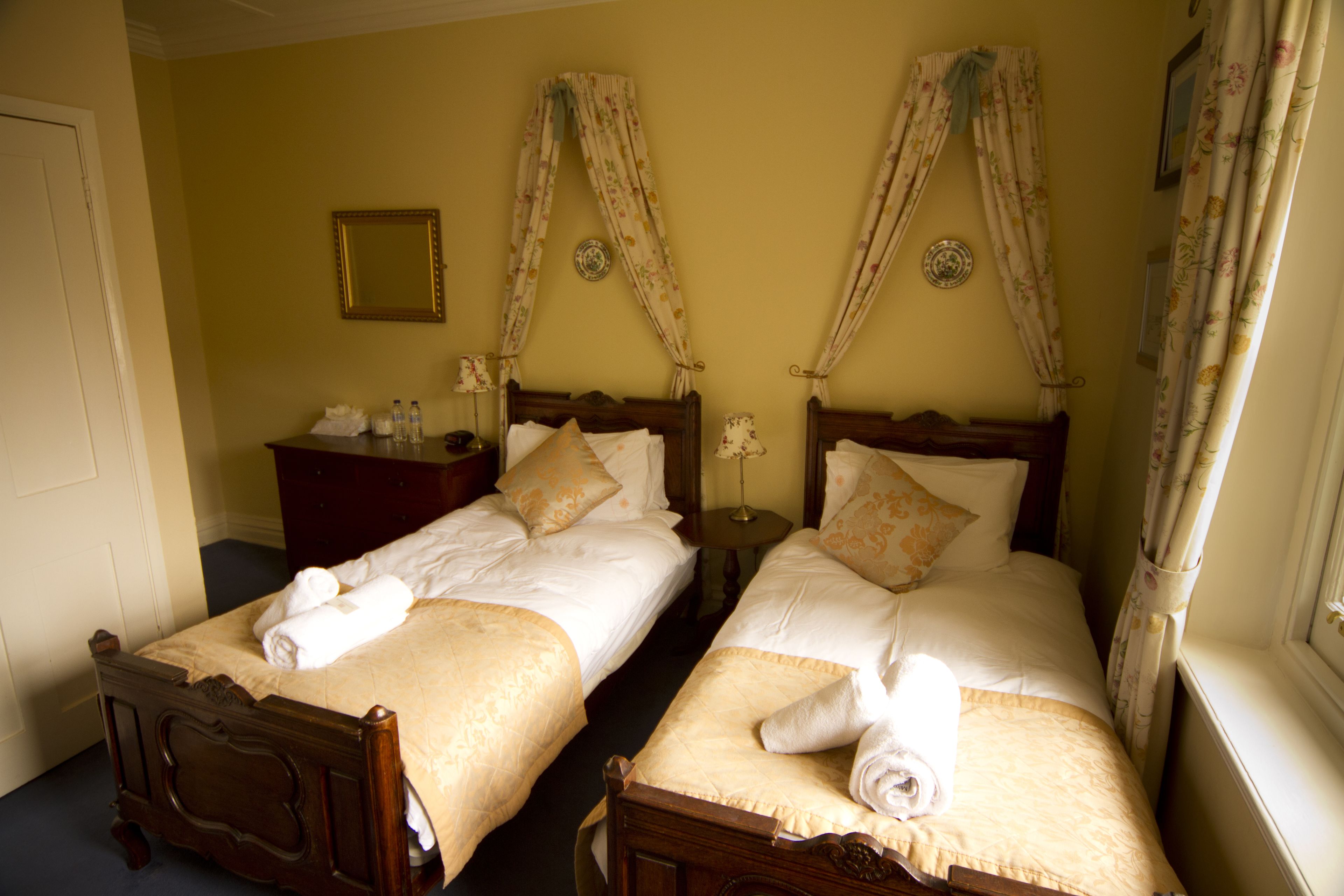 A hotel room with two wooden beds, each with a headboard, a bedside table, and a mirror on the wall.