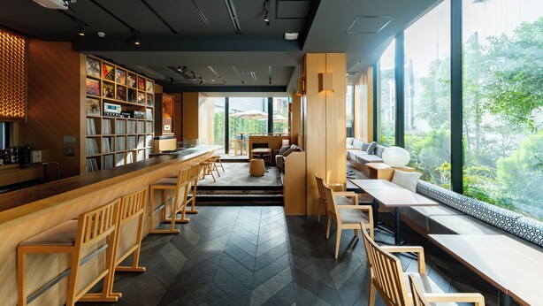 Uma área de bar moderna com mobiliário de madeira, uma estante de livros e grandes janelas com vista para o verde.