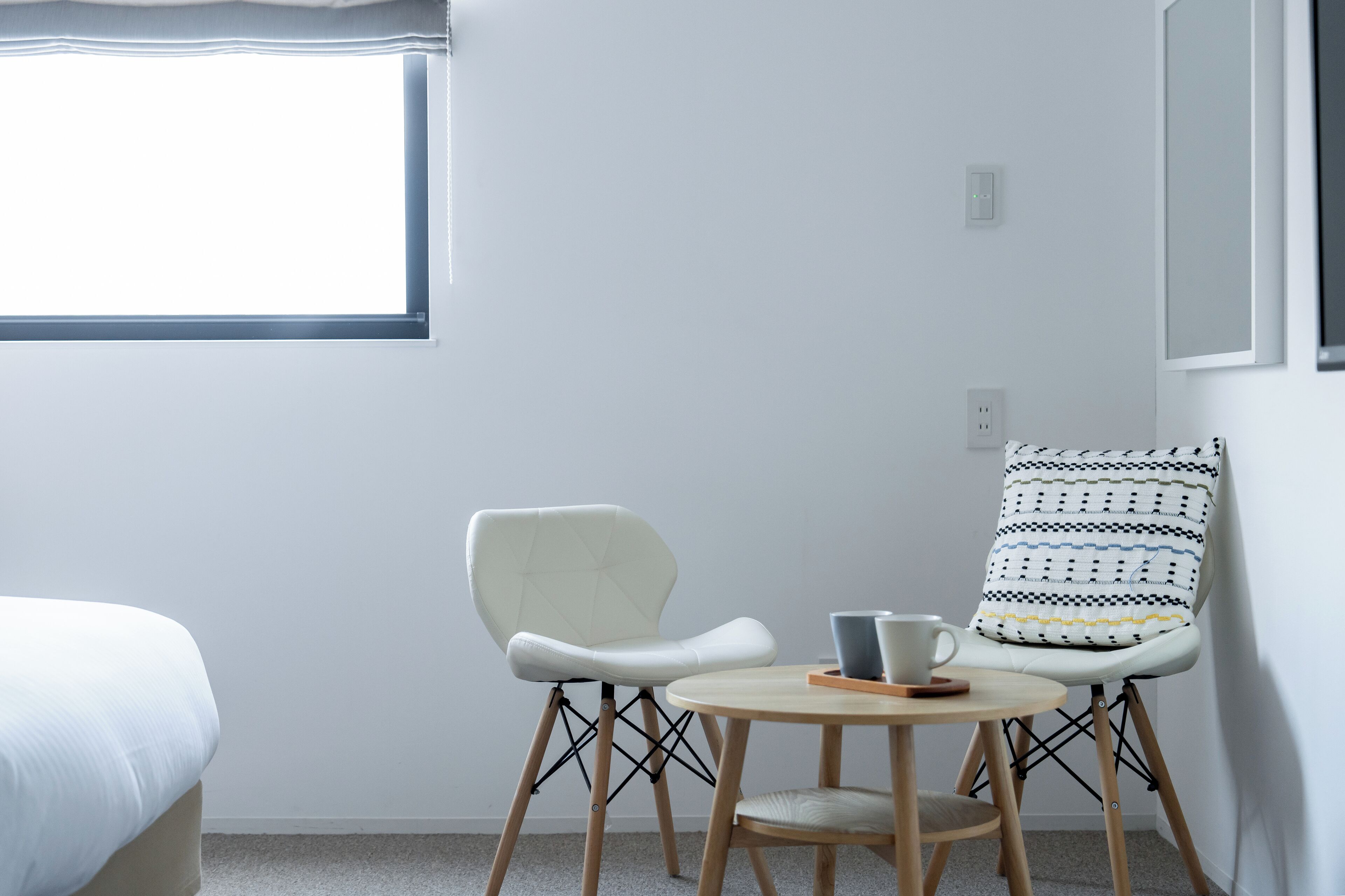 Una habitación minimalista con una cama, una ventana, una silla blanca con cojín estampado y una pequeña mesa redonda con una taza y platillo.