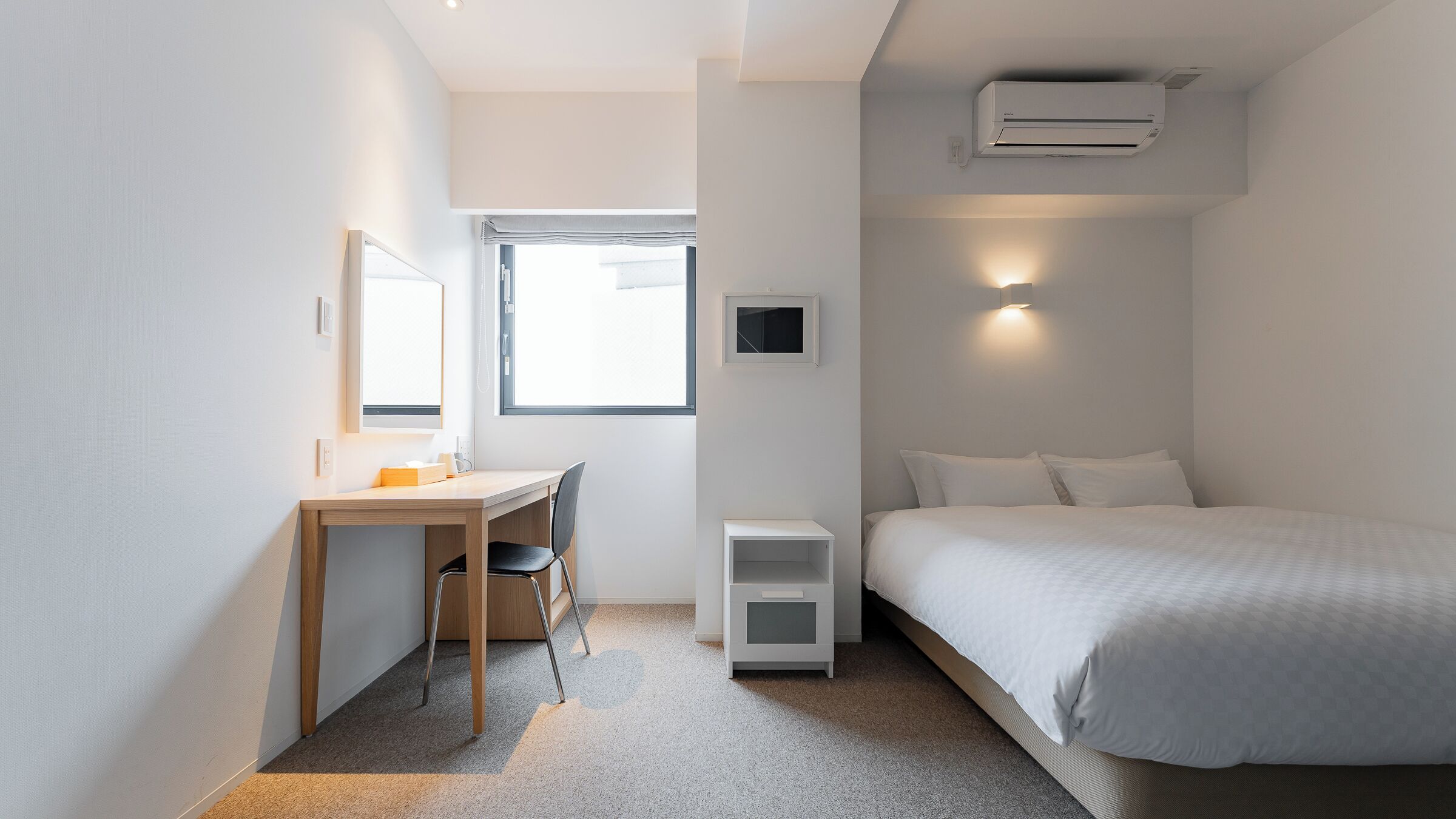 Een strak ingerichte hotelkamer met een bed, bureau, stoel, een klein nachtkastje, een wandairconditioner en een raam met uitzicht.