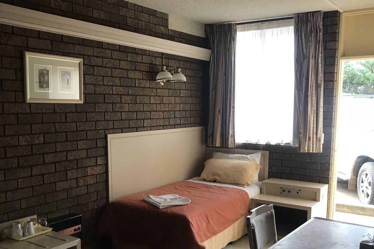 Een hotelkamer met een bakstenen muur, een bed met een rode sprei, een bureau met een stoel, een tafeltje en een raam met gordijnen.